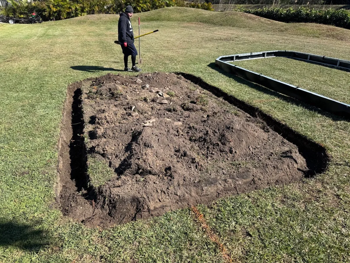A rectangle of lawn being dug out, a trench cut around its edge, with a worker holding a shovel beyond it and retaining wall panels lying on the grass