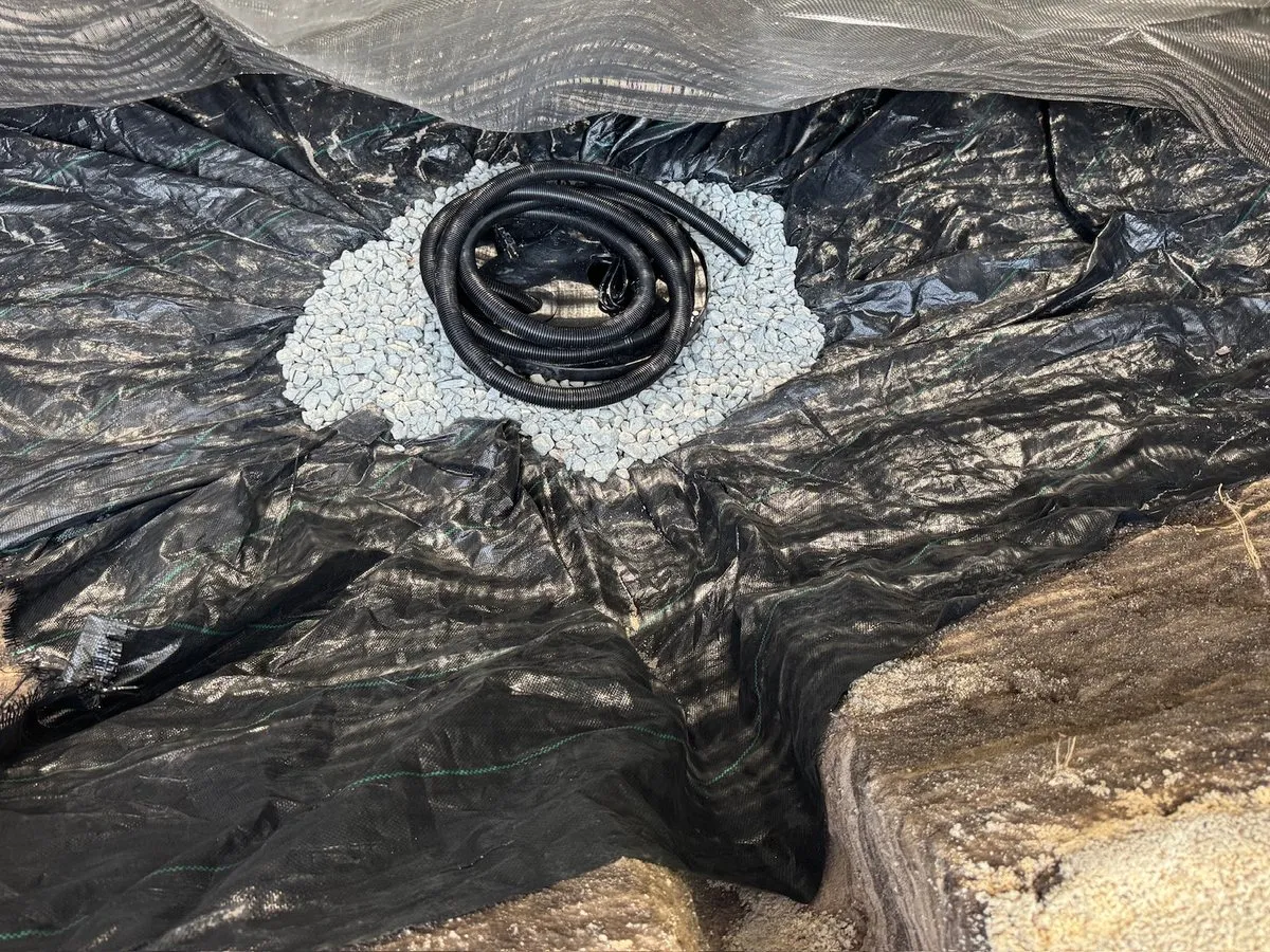Inside a lined pit: a coiled black hose resting in a patch of pale gravel on black fabric, with a sandy wall of the dig to one side
