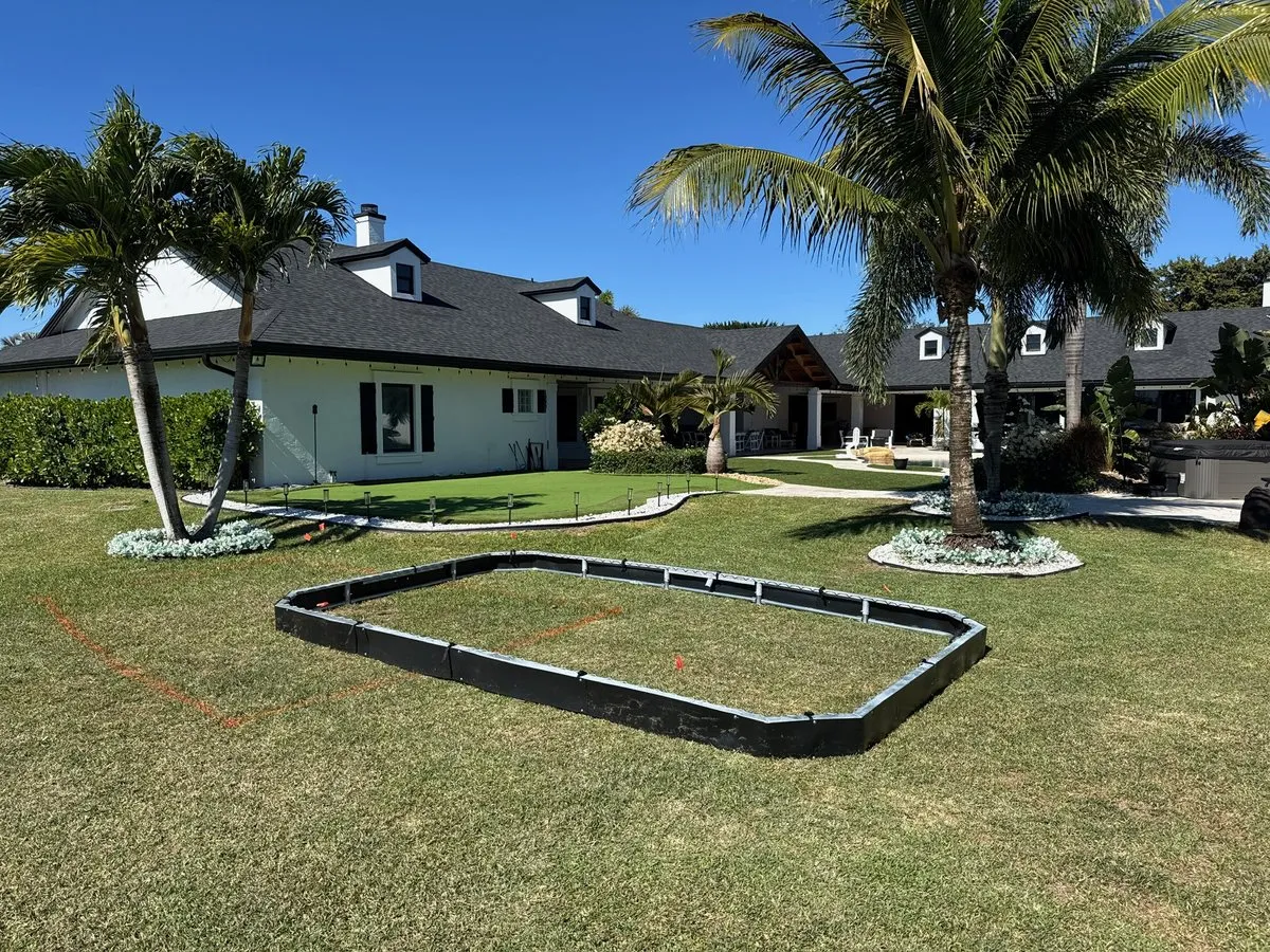 Black retaining wall panels joined into a rounded rectangle on a lawn, with orange marking paint, palms and a white house behind