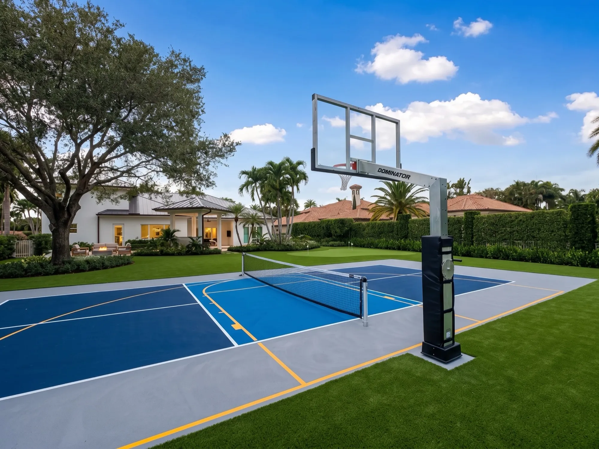 Blue and grey sport court marked for both basketball and pickleball, with a Dominator goal and a net across the middle, edged with artificial turf and a putting green beyond