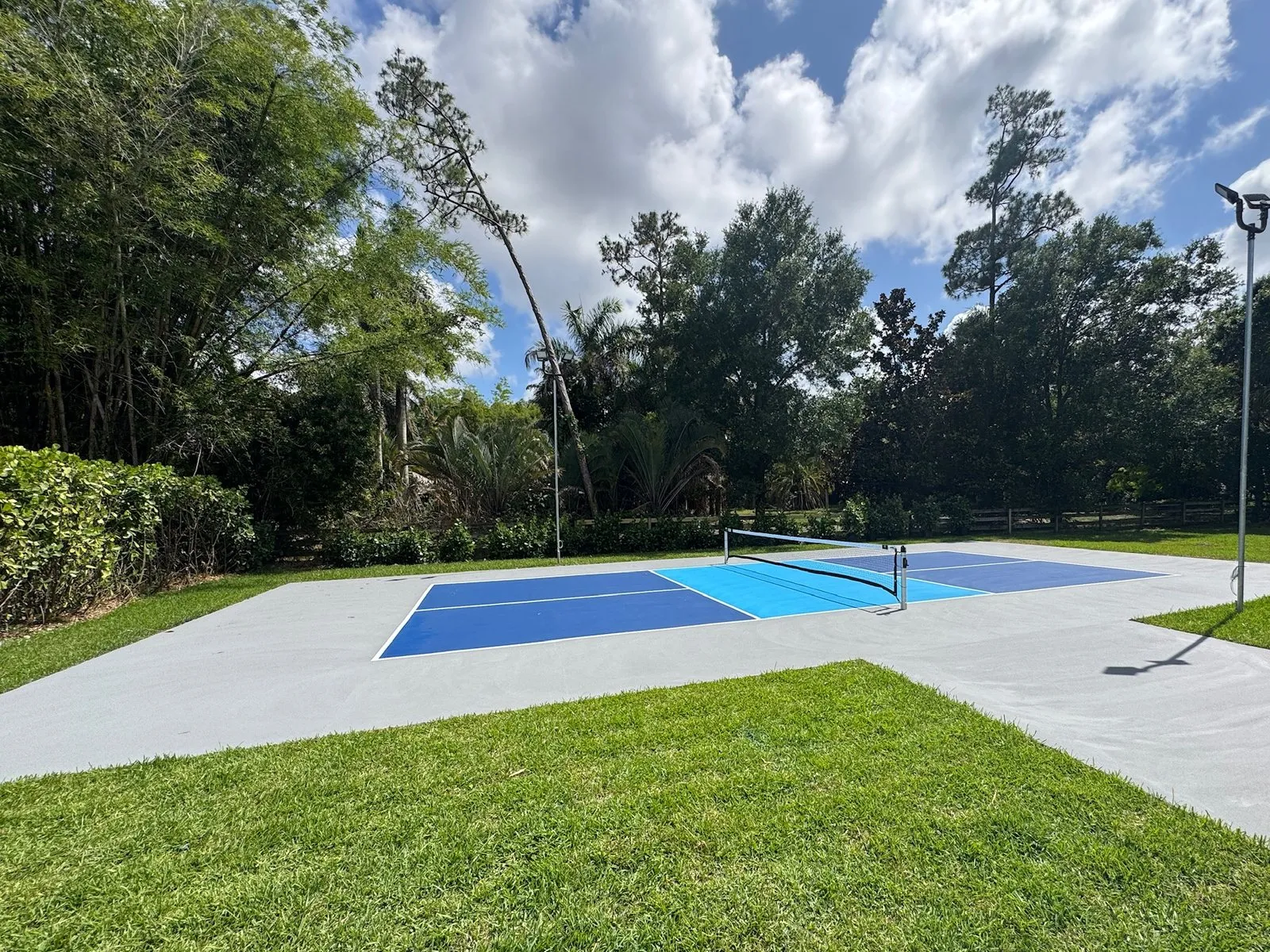 Pickleball court in two blues on a light grey slab with the net up, a court light on a pole to one side and a lawn in front