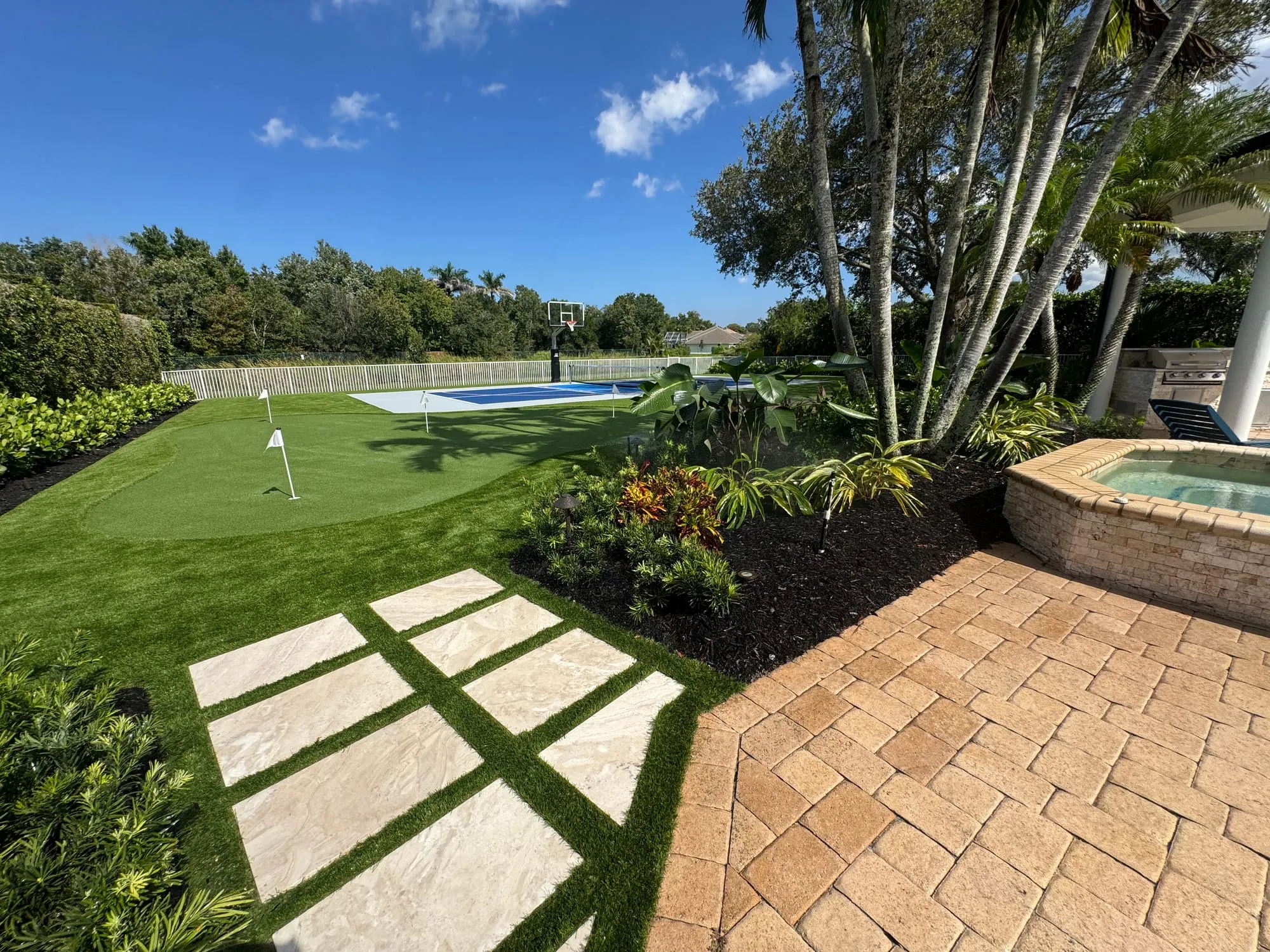 Travertine stepping stones set in artificial turf beside a brick paver deck and the corner of a spa, with a putting green and a blue sport court beyond.