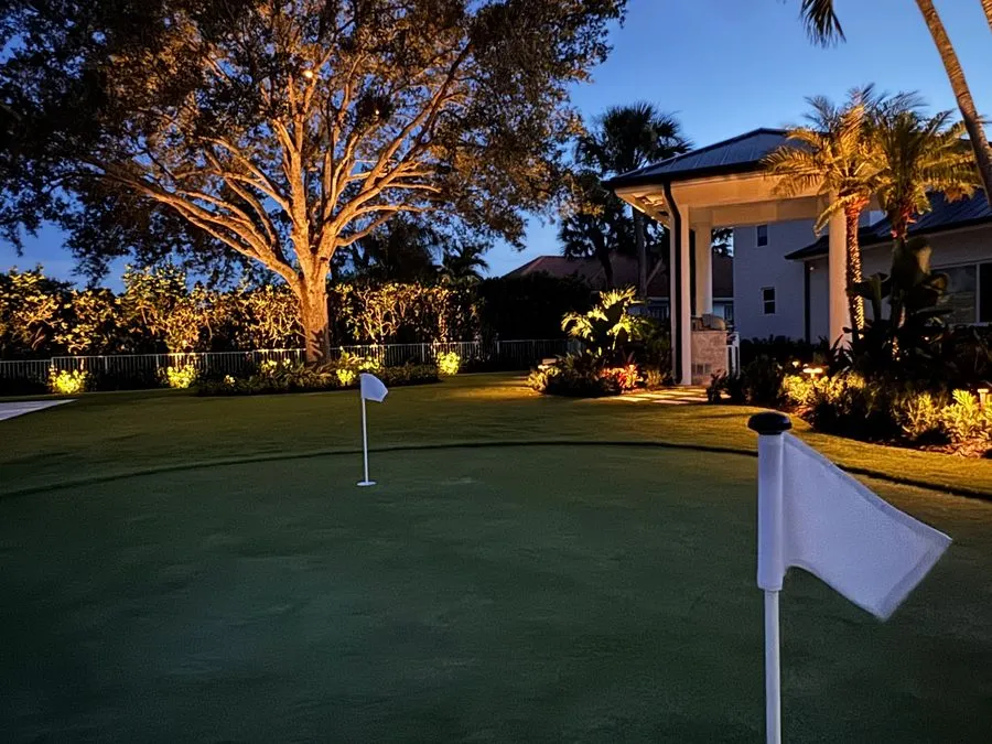 A putting green at dusk with a white flag close in the foreground and another in the far cup, an uplit oak and a white house with a covered porch behind.