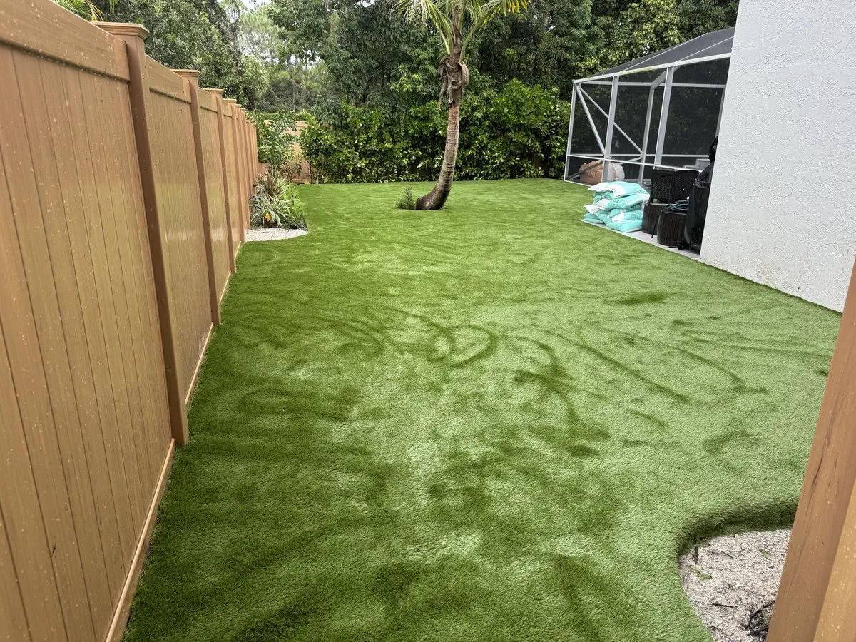 A tan vinyl privacy fence running the length of a side yard laid with artificial turf, with a palm tree and a screened pool enclosure at the far end