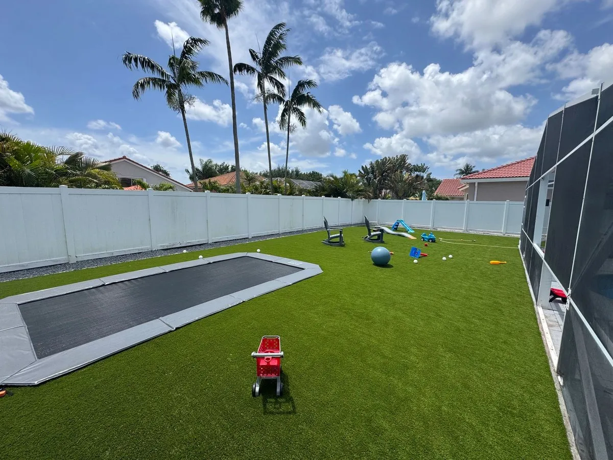 A long rectangular in-ground trampoline set flush in artificial turf, with a white vinyl fence and palm trees behind