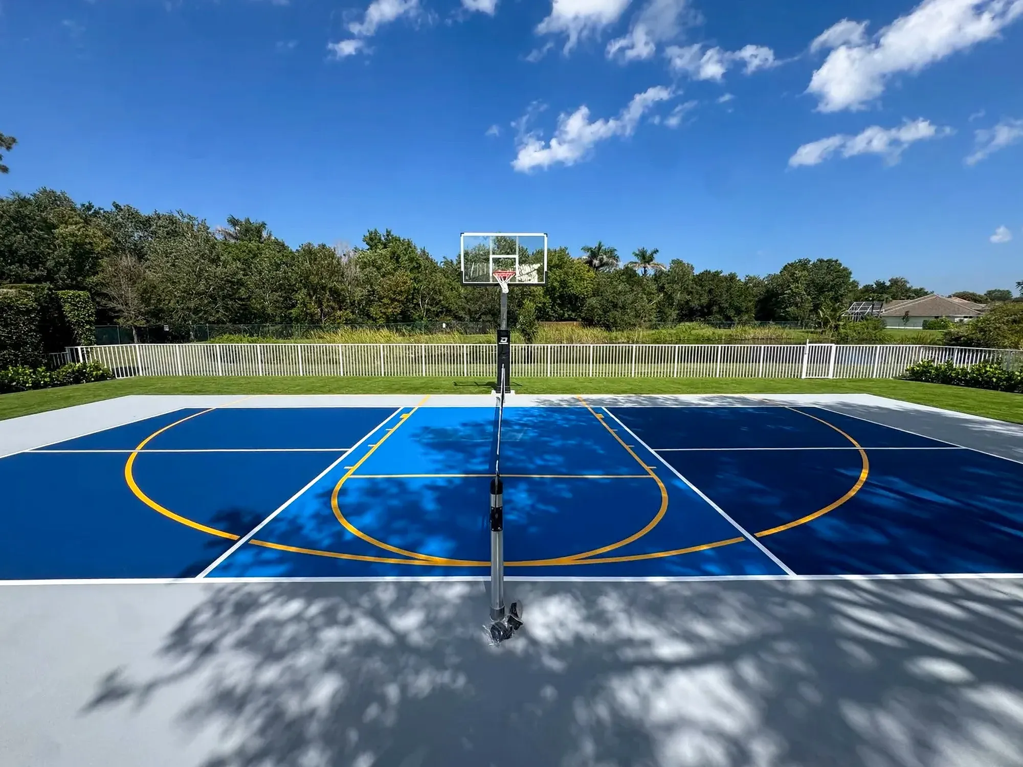 Backyard basketball court in two shades of blue with an adjustable glass goal, inside a white perimeter fence
