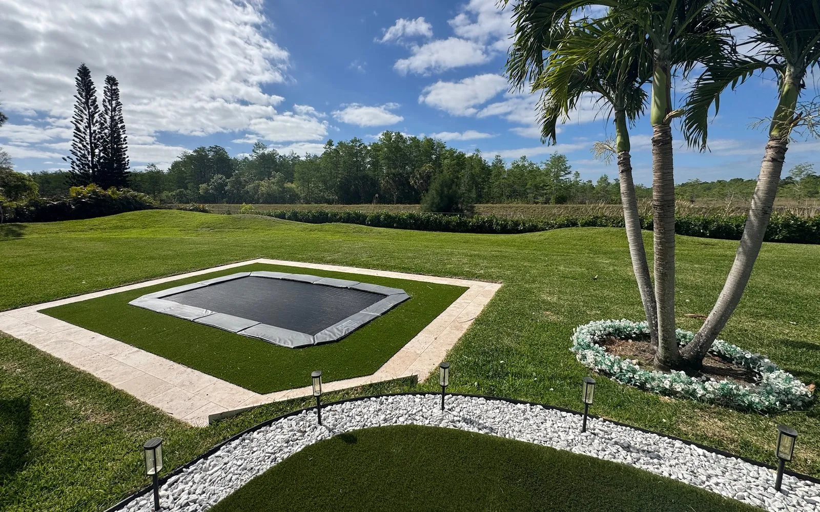 Rectangular in-ground trampoline set flush in artificial turf inside a pale paver border, with a palm beside it and a white stone path lined with path lights in front